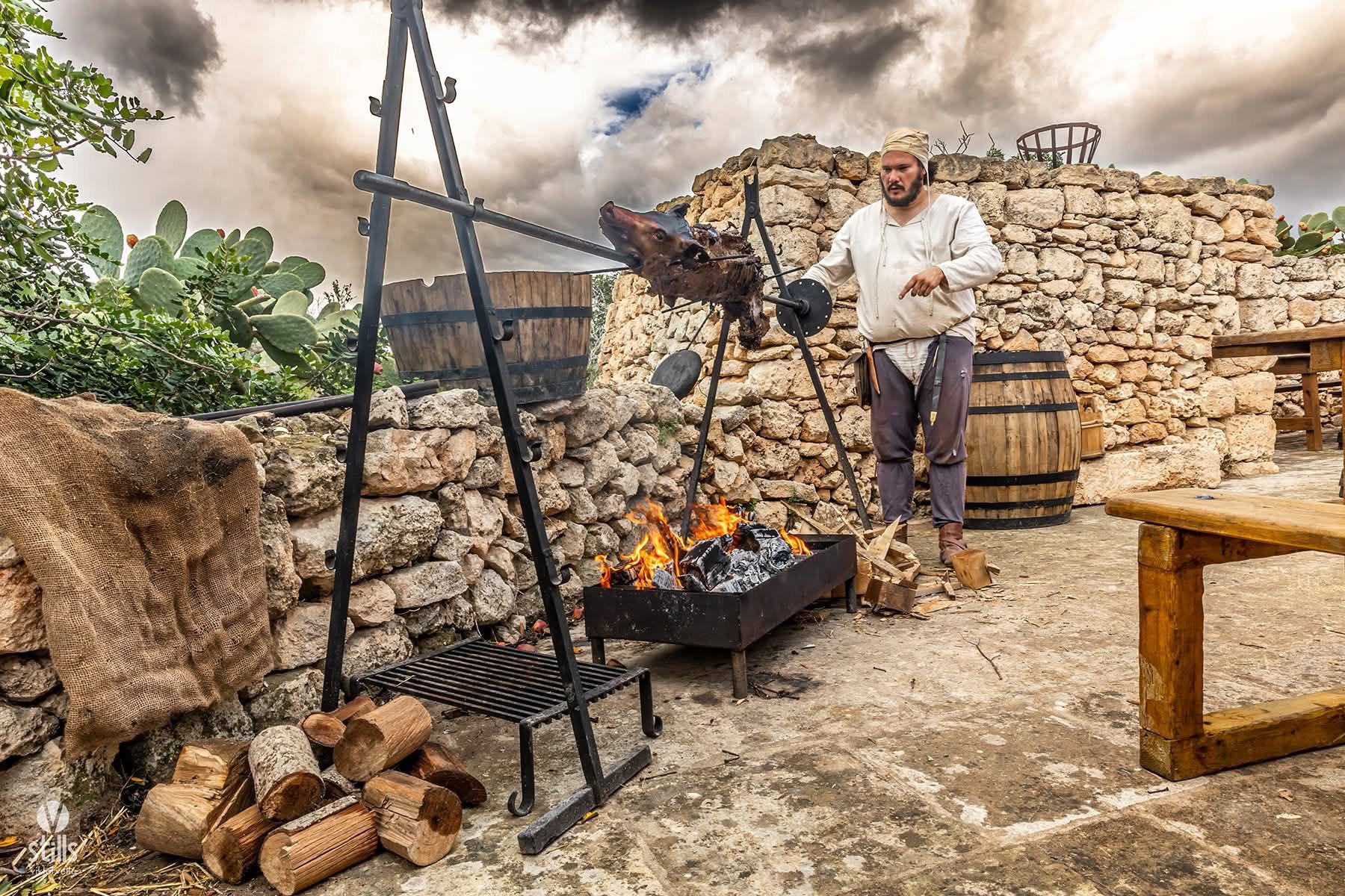 A participant of Anakron Living History tending meat roasting over an open fire during a medieval cooking reconstruction at the Selmun Interpretation Centre in Malta.